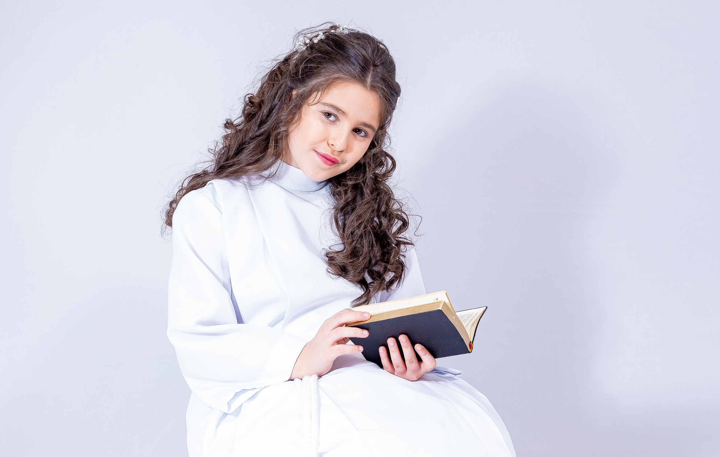 Jeune fille en aube blanche assise, lisant un livre, regard vers l’objectif