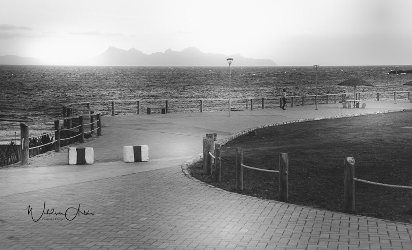 Promenade côtière minimaliste, silhouette solitaire, montagnes au loin