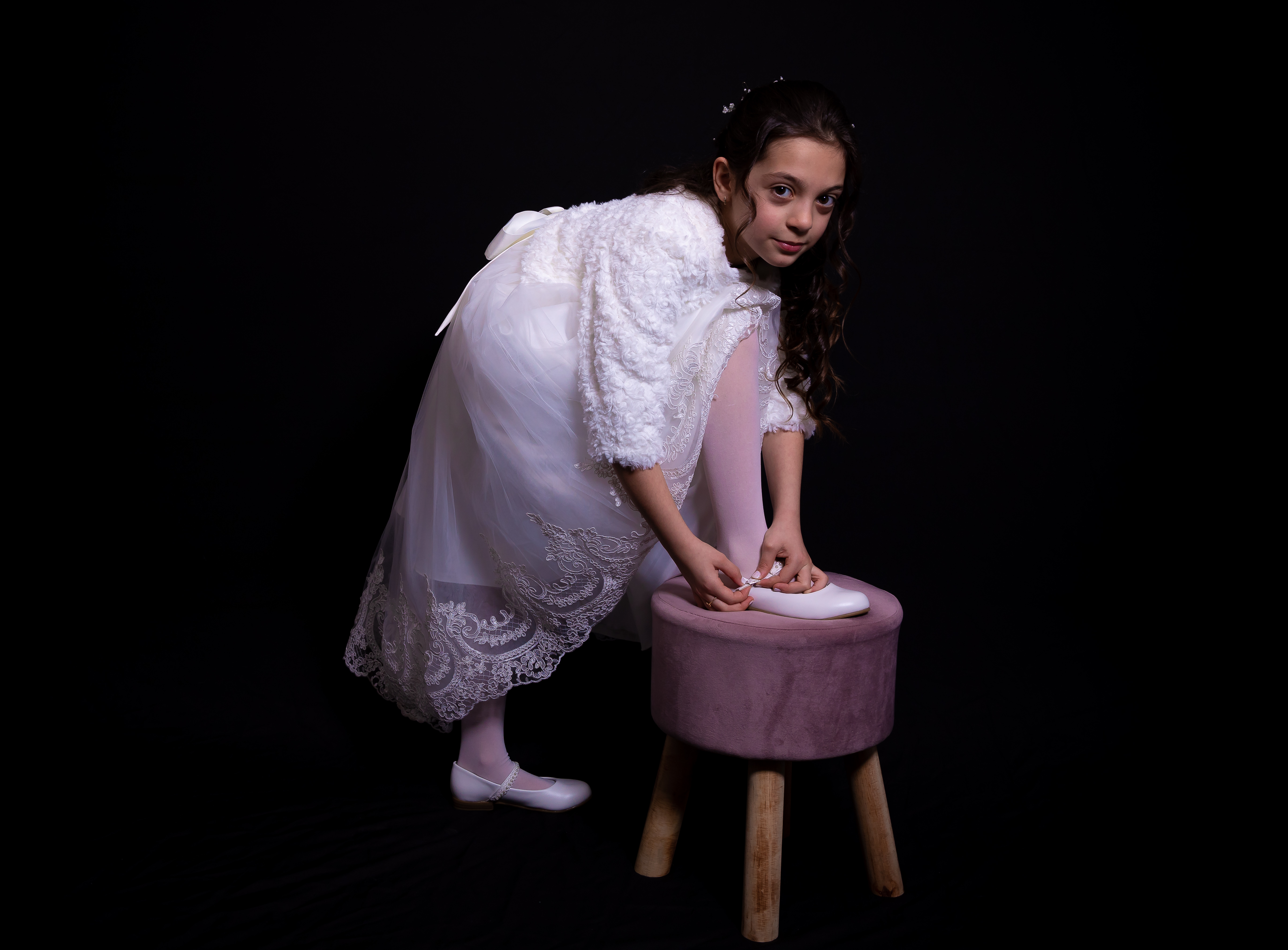 Jeune fille en robe de communion penchée, ajustant sa chaussure sur un tabouret