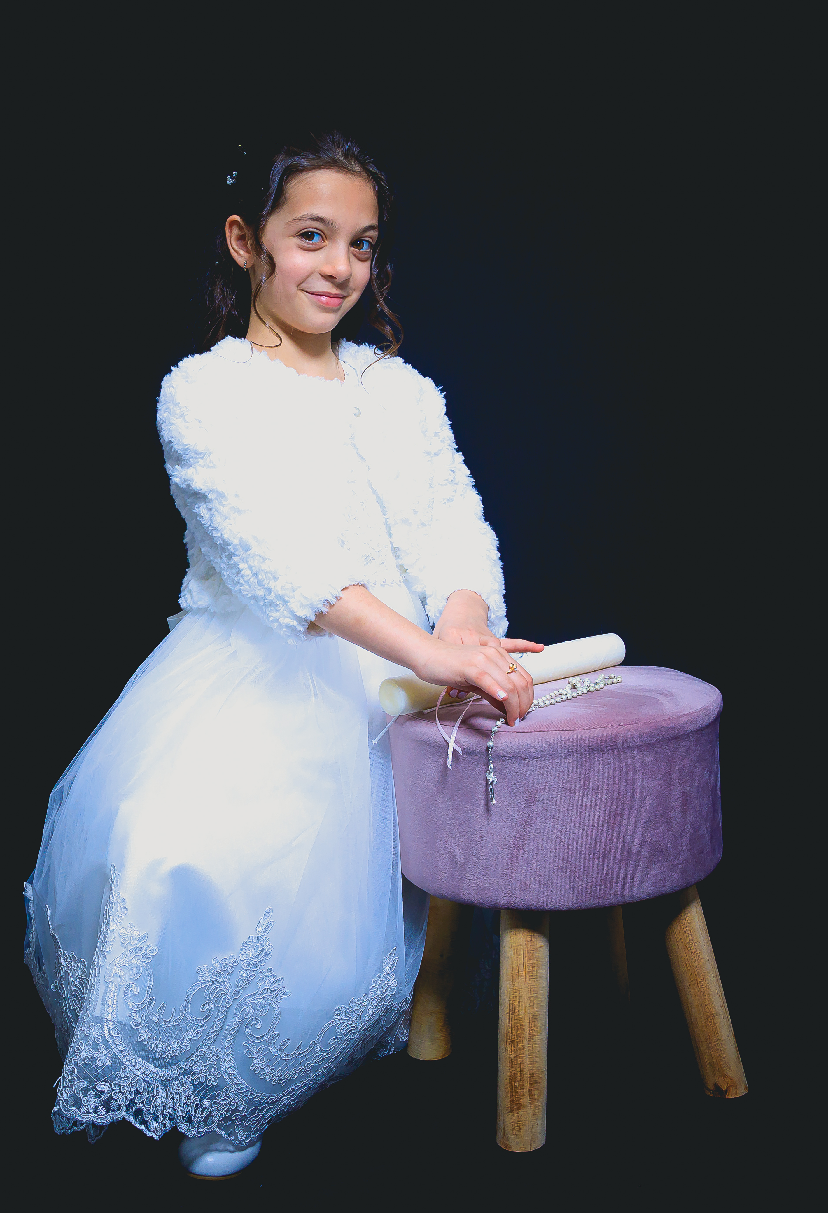 Jeune fille en robe de communion agenouillée près d’un tabouret, cierge et chapelet, sourire