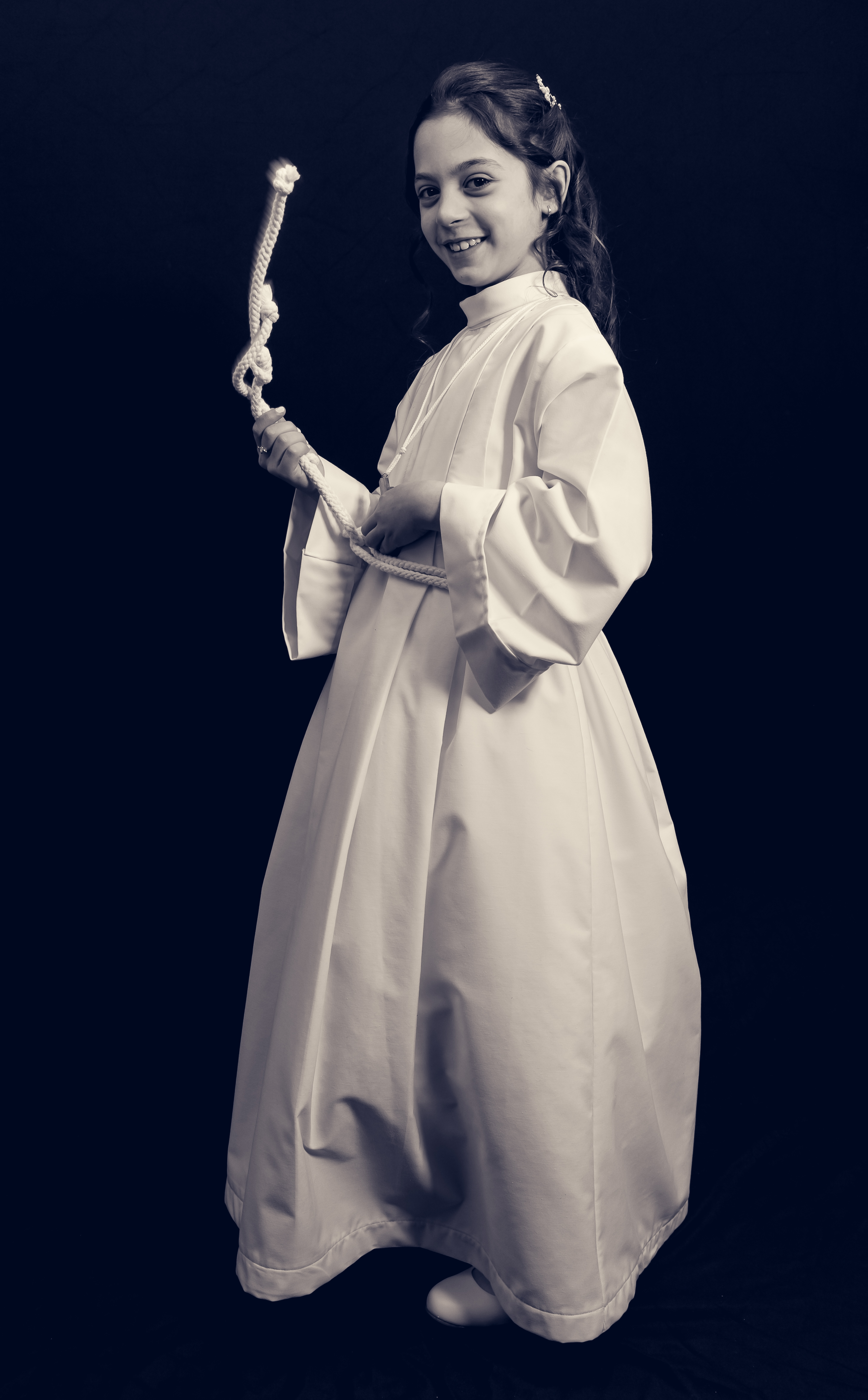 Jeune fille en aube de communion, vue de profil, tenant un cordon, sépia